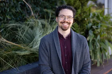 Photograph of Tyler, smiling, with short, curly hair, circular glasses and a goatee. He's wearing a gray cardigan over a maroon button up shirt, sitting on a bench in front of some foliage.