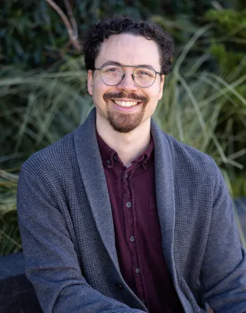 Closeup photograph of Tyler, smiling wearing a gray cardigan over a maroon button up shirt, sitting on a bench in front of some foliage.